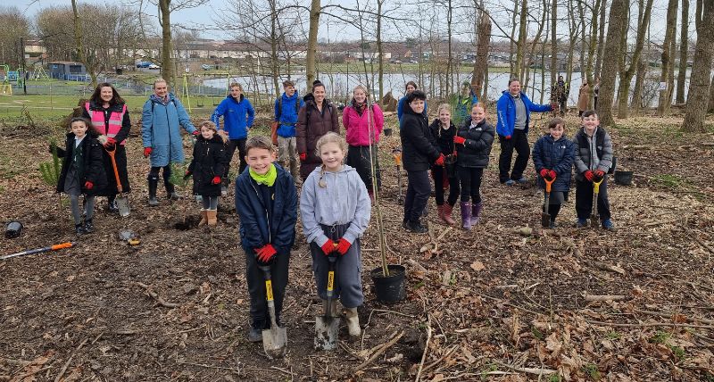 A group of children with shovels and spades and saplings in pots in a woodland with a lake behind